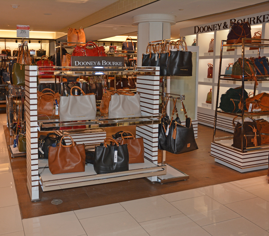 gallery image of custom store-in-store retail interior project featuring secure polished stainless steel T-stands and display shelving, retractable cable-secured handbag displays, and custom logos and signage fabricated and installed for dooney & bourke by Visual  Millwork